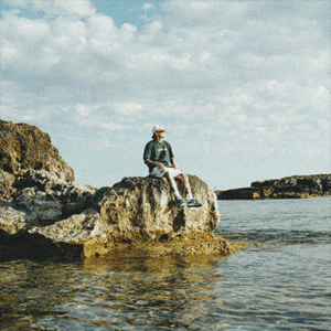 Estikay sitzt auf einem großen Stein, blickt in die Ferne; um ihn herum Wasser, im Hintergrund Felsen