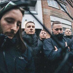 Die Band Malevolence steht vor einem Haus und schaut durch einen Maschendrahtzaun. Sie wirken nachdenklich.