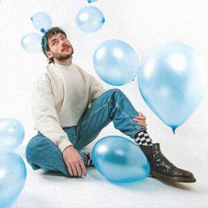 James Marriott sitzt auf dem Boden und schaut nach oben. Hellblaue Luftballons fallen von oben runter.