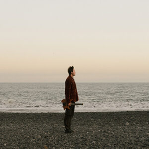 Plini steht am Meer, seitlich gedreht, Blick zur Seite