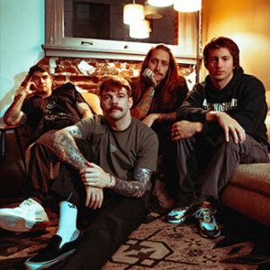 Bandfoto von The Home Team. Die Band sitzt zusammen auf einer Couch und davor auf dem Boden