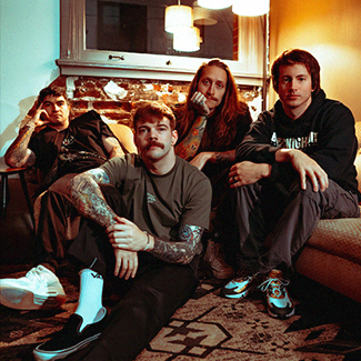Bandfoto von The Home Team. Die Band sitzt zusammen auf einer Couch und davor auf dem Boden